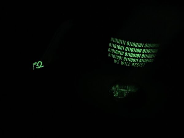 Image showing two Slash-32 snapback hats in dark, highlighting glow-in-the-dark binary patch with “We Will Resist” on one hat and glowing /32 microchip patch on the other.