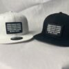 Front view image showing two Slash-32 snapbacks, black and white, both with black rectangular patch with binary and “We Will Resist” text.