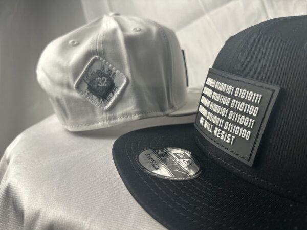 Image featuring a black Slash-32 9/50 Snapback hat with a black binary patch and a white fitted hat with a white diamond patch, both showcasing the /32 microchip emblem.