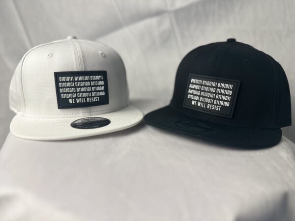 Front view image showing two Slash-32 snapbacks, black and white, both with black rectangular patch with binary and “We Will Resist” text.
