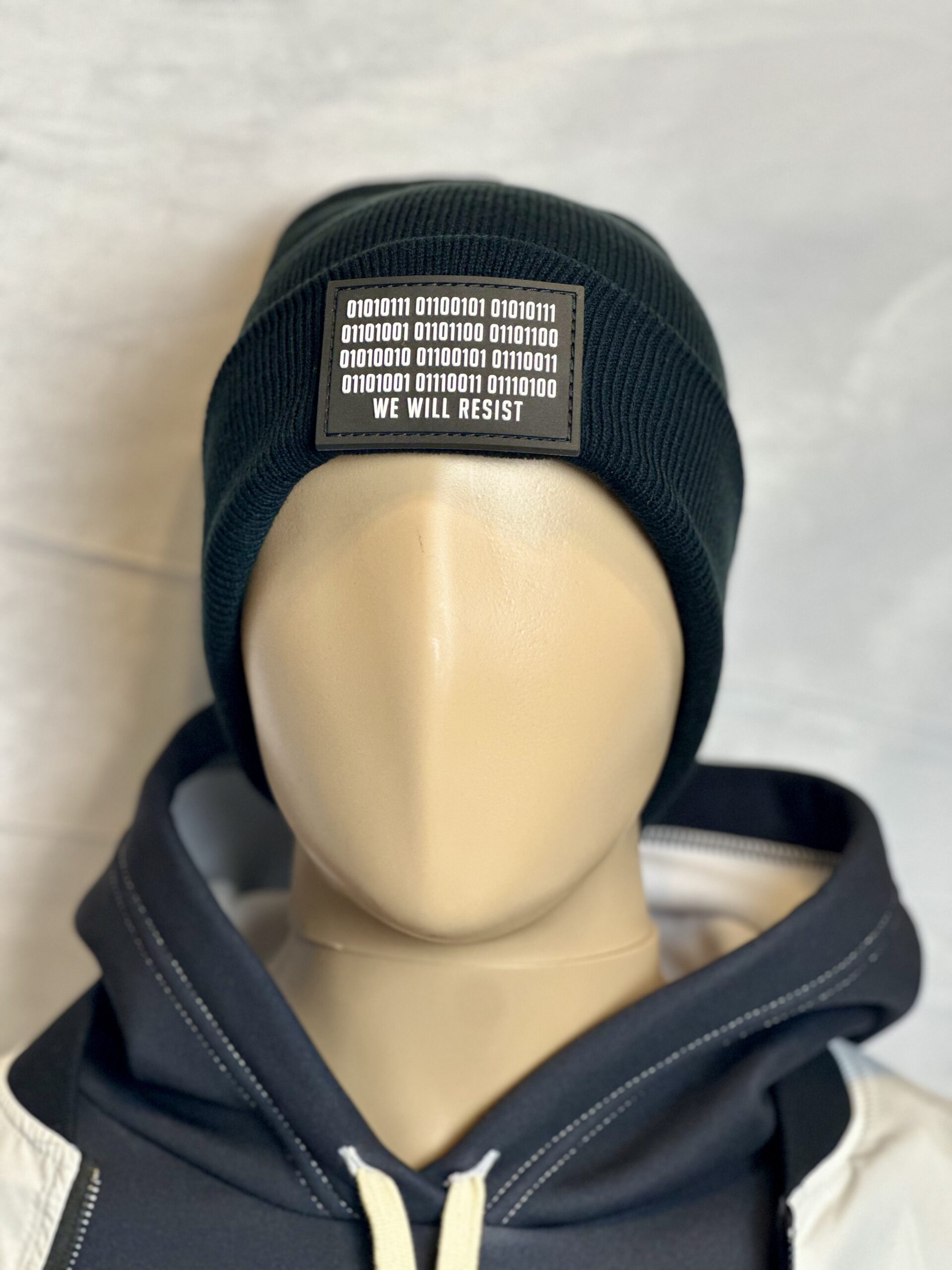 Image of the black “We Will Resist” rolled beanie by Slash-32 on a mannequin wearing a hoodie, featuring a grey rectangle patch with binary matrix and “We Will Resist” text.