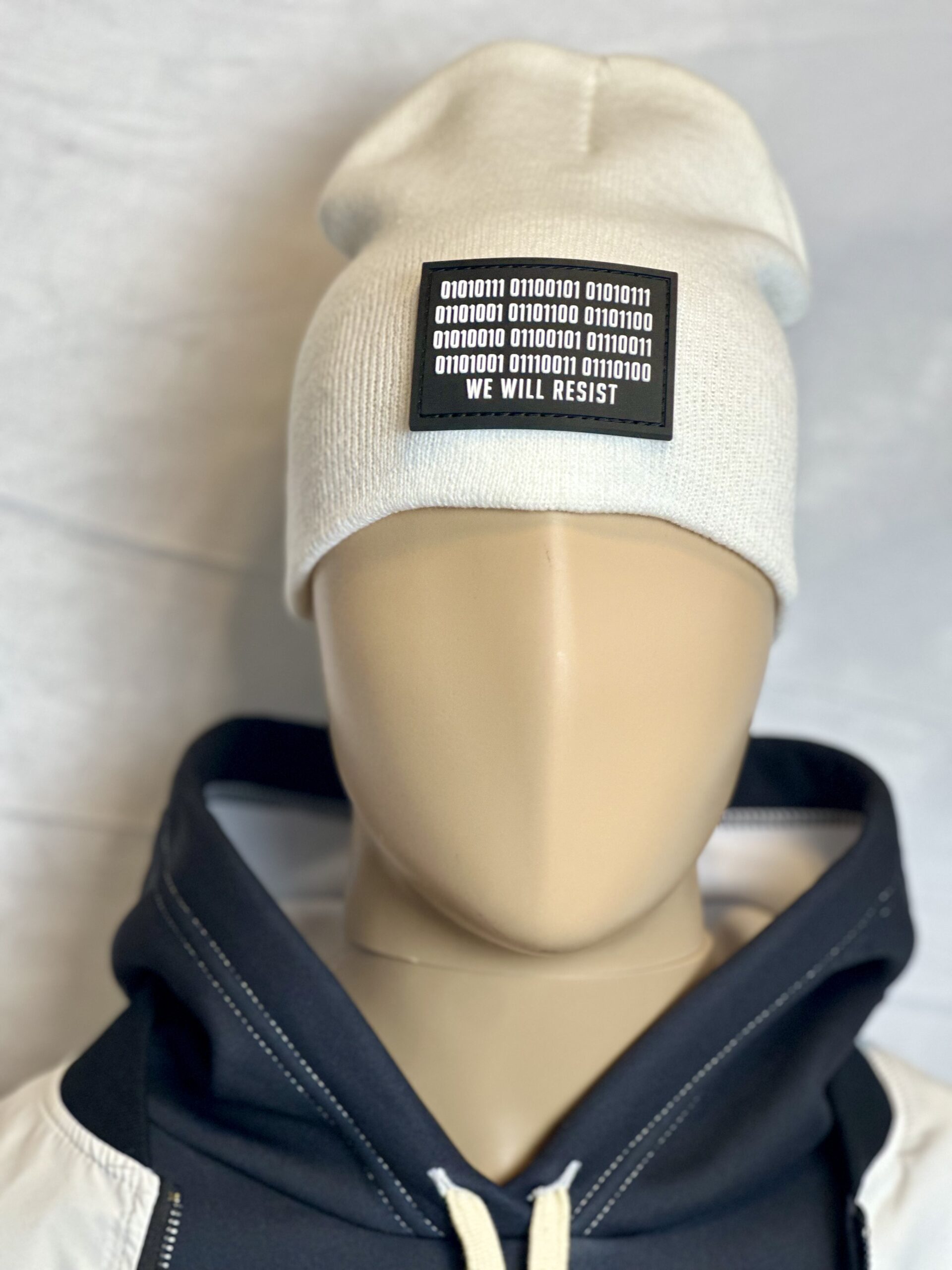 Image of white slouch style fitted beanie on mannequin with rectangular black patch featuring binary code above “We Will Resist” text, sewn onto front of hat.