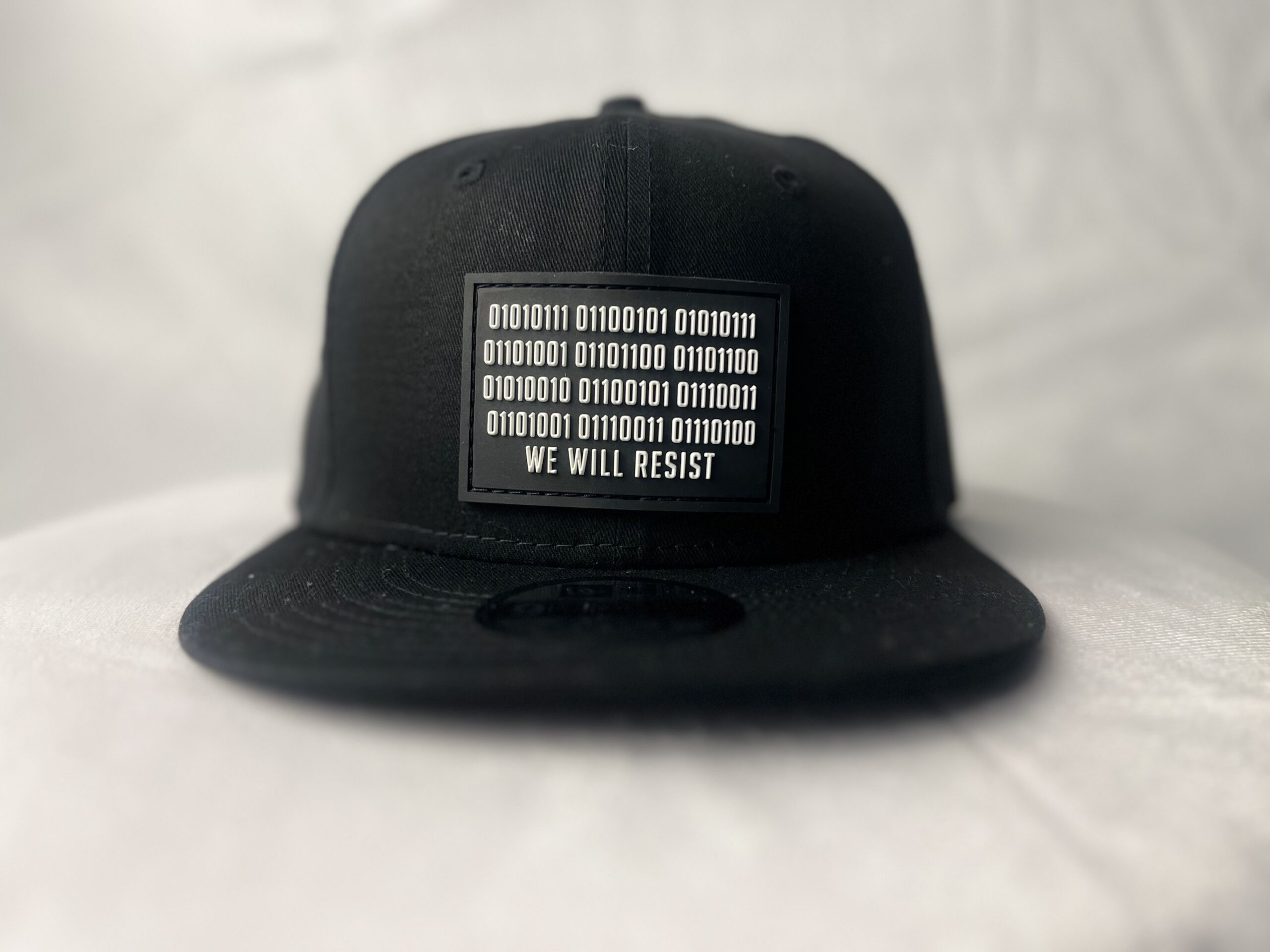Front view of the Slash-32 9/50 Snapback hat, Black (Panda Mode) "We Will Resist" cap, displaying a black rectangle patch with binary code above the “We Will Resist” text.