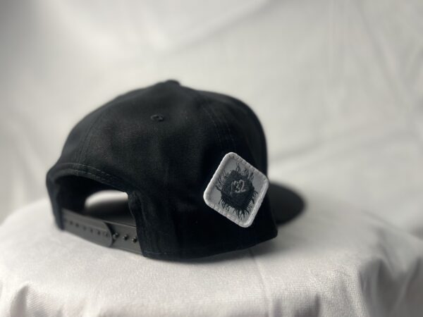 Side-back view of the Slash-32 9/50 Snapback hat, Black (Panda Mode) "We Will Resist" cap, featuring a white diamond-shaped patch with the /32 microchip emblem stitched in.