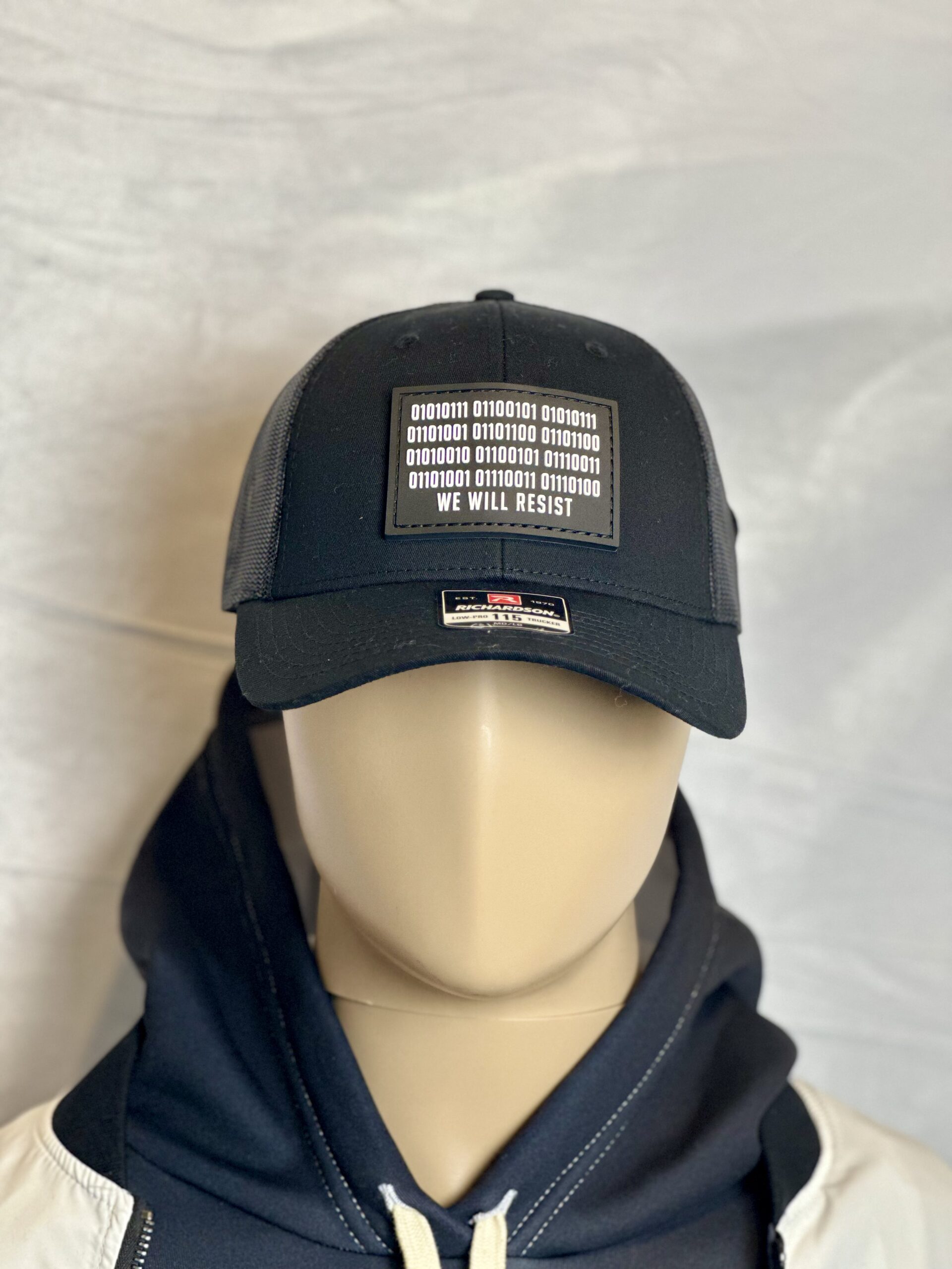 Image of Slash-32 Trucker Style Snapback hat - Black "We Will Resist" cap on mannequin, featuring a black rectangle patch with binary matrix and “We Will Resist” text on the front.