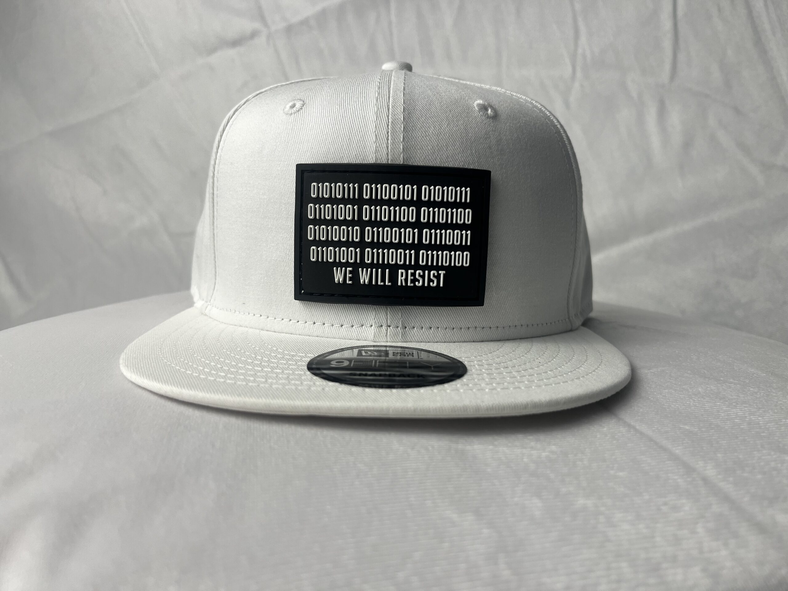 Image of Slash-32 9/50 Snapback hat, White showing the black rectangular patch embroidered with binary code and “We Will Resist” text on the front.