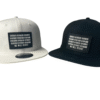 Front view image showing two Slash-32 snapbacks, black and white, both with black rectangular patch with binary and “We Will Resist” text.