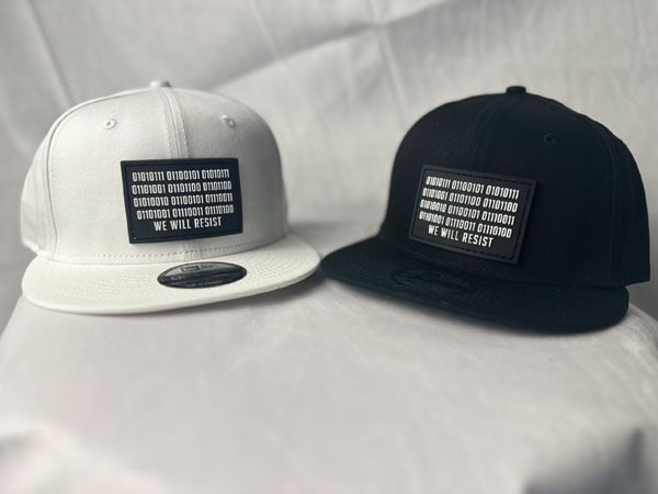 Front view of two Slash-32 9/50 Snapback hats—white and black—both featuring rectangular patches embroidered with binary code and the text “We Will Resist.”